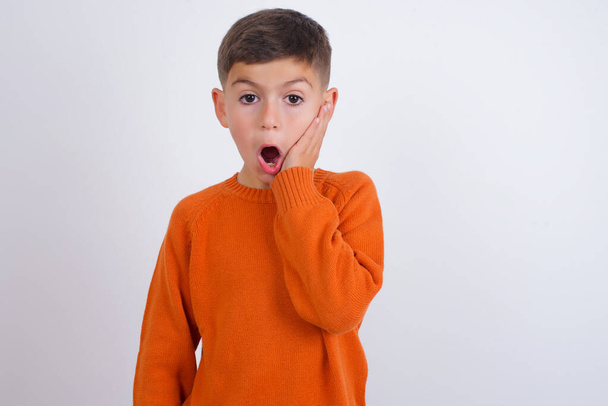Schockierter kaukasischer Junge in gestricktem orangefarbenem Pullover, der vor weißem Hintergrund steht, schaut mit großer Überraschung sehr fassungslos, erstaunt über unerwartete Neuigkeiten, Gesichtsausdruck Konzept. - Foto, Bild