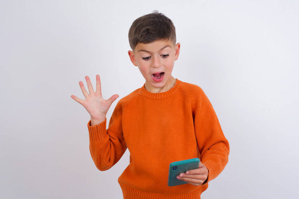 Caucasian kid boy wearing knitted orange sweater standing over white background holding in hands cell reading browsing news - Photo, Image