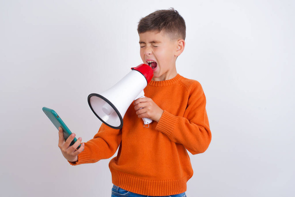 Bláznivý dojem kavkazský chlapec nosí pletený oranžový svetr stojící nad bílým pozadím pomocí mobilního telefonu čte neuvěřitelné novinky na sociálních médiích informace zírat, křičí wow omg. - Fotografie, Obrázek