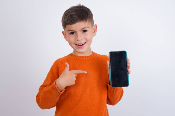 白い背景の上に立ってニットオレンジセーターを着て笑顔白人の子供の少年は、コピースペースをモックアップします。空白の画面を持つ携帯電話上のインデックス指を指す - 写真・画像