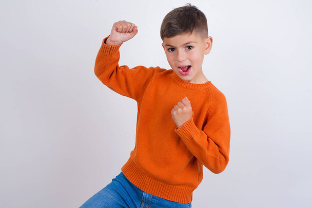 Photo de profil de garçon caucasien excité portant un pull tricoté orange debout sur fond blanc levant les poings célébrant le vendredi noir shopping - Photo, image