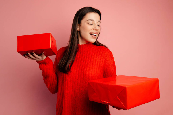 Schöne junge Frau zeigt ihre Geschenke in der roten Verpackung und lächelt. Das Mädchen trägt glattes schwarzes Haar, roten Pullover und schwarze Maniküre - Foto, Bild