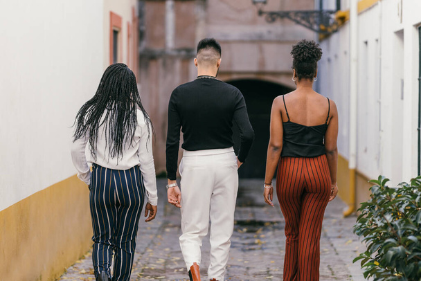 Back of a man and two women in modern clothes walking through a street - Фото, изображение