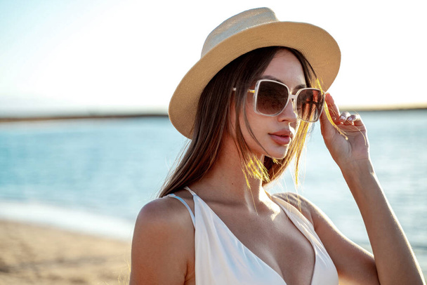 Young beautiful woman enjoying summer vacation, walking at the beach. Portrait of brunette woman with long hair wearing fashionable sunglasses and hat. Tourism. Real people lifestyle - Fotografie, Obrázek