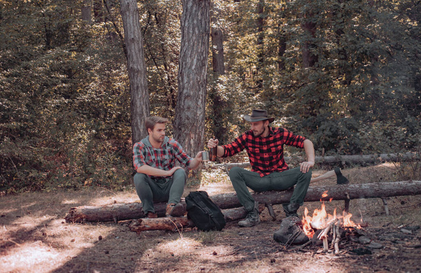 Vacation weekend picnic camping and hiking. Two young guy sitting in the forest and drinking beer near campfire. Friends spend leisure weekend forest nature background. - Fotografie, Obrázek