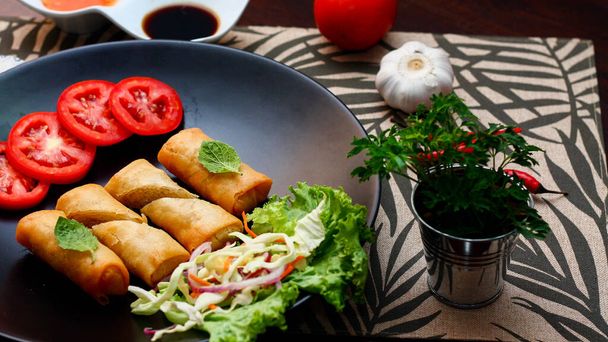 Fried spring rolls with vegetables and tomatoes placed in a black plate on a black wooden table and dipping sauce in tjhailand. - Photo, Image