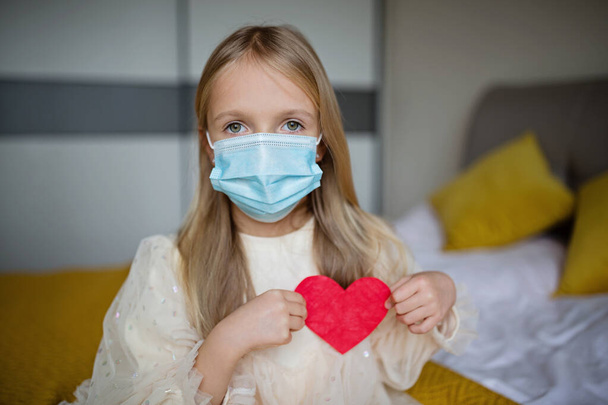 Little school kid girl holding red heart. Valentines day during pandemic coronavirus covid-19 quarantine. Child showing heart online via video chat to boy friend - Foto, Imagen