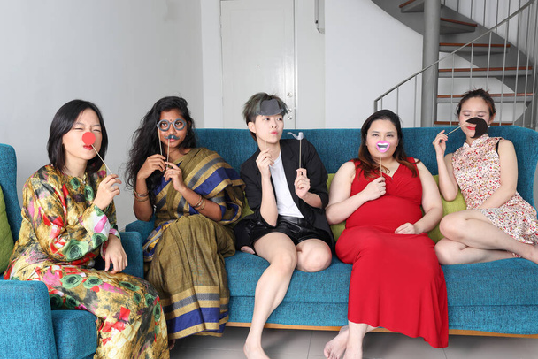 Joven mujer asiática grupo charla cantar haciendo divertido burlas caras expresión selfie retrato papel accesorios bigote sombrero tubería nariz pelo en azul sala de estar sofá sentarse - Foto, Imagen