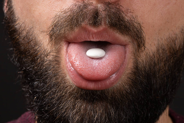 white round tablet on tongue of open mouth of bearded man with dark mustache on black background, take vitamins, painkiller - Photo, image