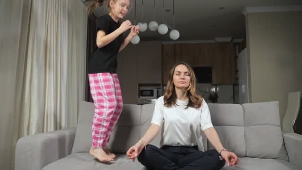 madre hace yoga mientras hija distrae saltar en sofá - Imágenes, Vídeo