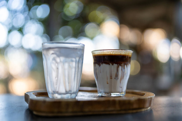 Грязный кофе или стакан эспрессо, смешанный с холодным свежим молочным слоем, подается с водой на деревянном подносе на столе. Кофейня кофе меню. - Фото, изображение