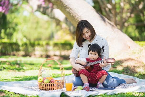 Çocuklu bir aile bahar bahçesinde pikniğin tadını çıkarıyor. Ebeveynler ve çocuklar yaz parkında dışarıda öğle yemeği yerken eğleniyorlar. Anne ve kızı bahçede ukulele oynuyor. - Fotoğraf, Görsel