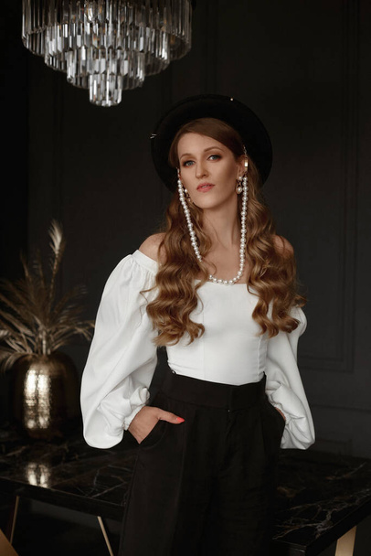 Mujer joven de moda en blusa blanca y sombrero negro de moda en el interior minimalista oscuro - Foto, Imagen