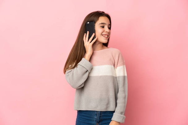 Niña aislada sobre fondo rosa manteniendo una conversación con el teléfono móvil con alguien - Foto, Imagen