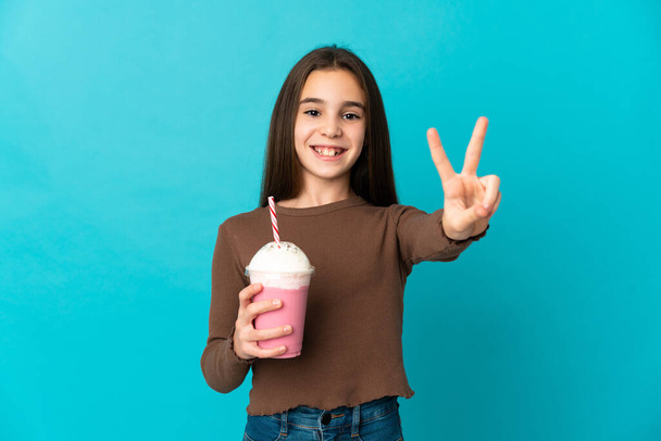 Kislány eper tejturmix elszigetelt kék háttér mosolygós és mutatja győzelem jele - Fotó, kép