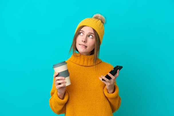 Mujer inglesa joven con chaqueta de invierno aislada sobre fondo azul sosteniendo café para llevar y un móvil mientras piensa algo - Foto, Imagen