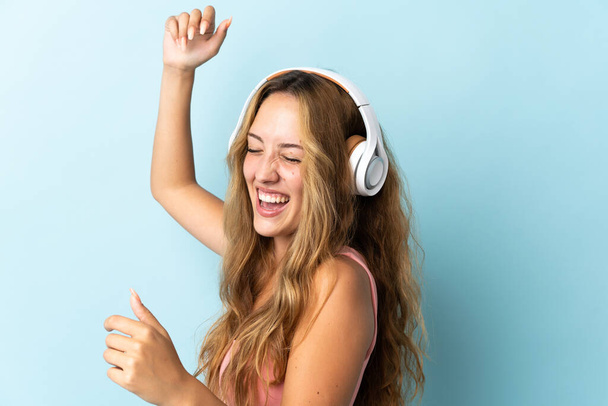 Νεαρή ξανθιά γυναίκα απομονωμένη σε μπλε φόντο ακούγοντας μουσική και χορεύοντας - Φωτογραφία, εικόνα