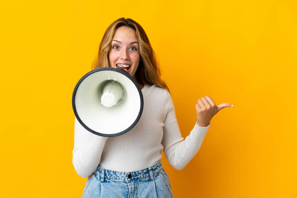 Young blonde woman isolated on yellow background shouting through a megaphone and pointing side - Fotoğraf, Görsel
