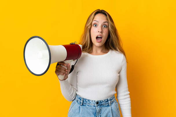 Young blonde woman isolated on yellow background holding a megaphone and with surprise expression - Φωτογραφία, εικόνα