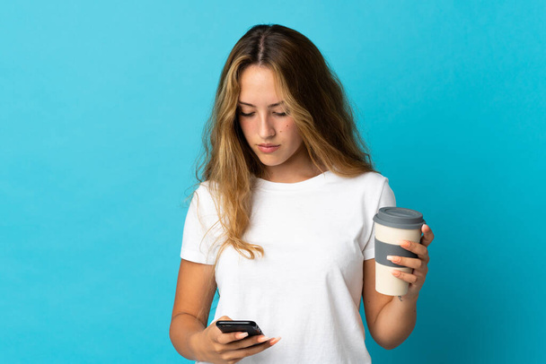 Nuori blondi nainen eristetty sinisellä taustalla tilalla kahvia ottaa pois ja mobiili - Valokuva, kuva