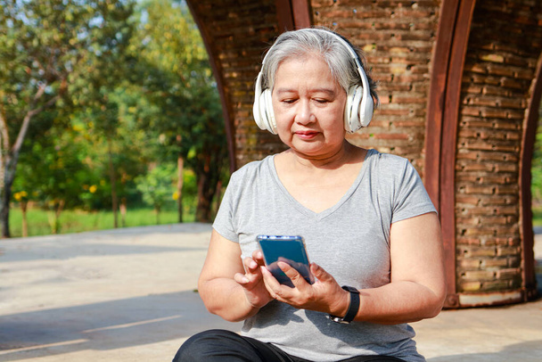 Azjatycka starsza kobieta ćwiczy rano w parku Siedzi i relaksuje się, trzymając smartfona i nosząc słuchawki do słuchania muzyki. pojęcie opieki zdrowotnej w podeszłym wieku - Zdjęcie, obraz