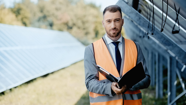 businessman in safety vest and suit holding folder near solar panels - Foto, afbeelding