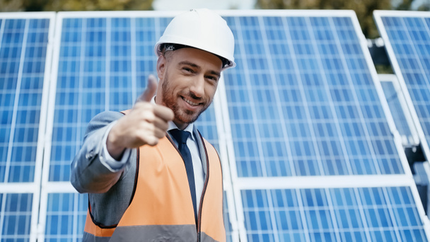 szczęśliwy biznesmen w hardhat pokazuje kciuk w górę w pobliżu baterii słonecznych  - Zdjęcie, obraz