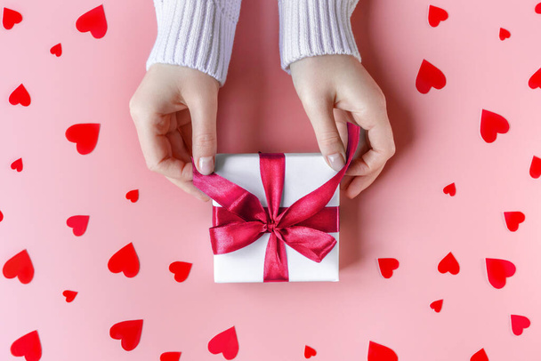 Female Hands holding a gift in white wrapping paper on pink background with red hearts - Zdjęcie, obraz