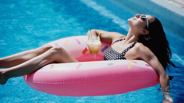 mujer complacida en gafas de sol sosteniendo vidrio con cóctel y nadando en anillo inflable en la piscina  - Foto, Imagen