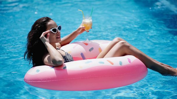 довольная женщина регулирует солнечные очки, держа стакан с коктейлем и плавая на надувном кольце в бассейне - Фото, изображение
