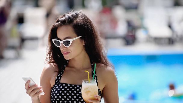 весела жінка в сонцезахисних окулярах і купальнику тримає коктейль і використовує смартфон
 - Фото, зображення