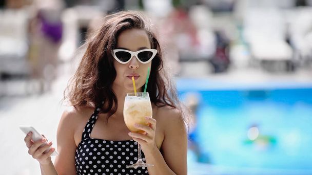 brunette woman in sunglasses and swimsuit drinking cocktail and holding smartphone - Foto, Imagem