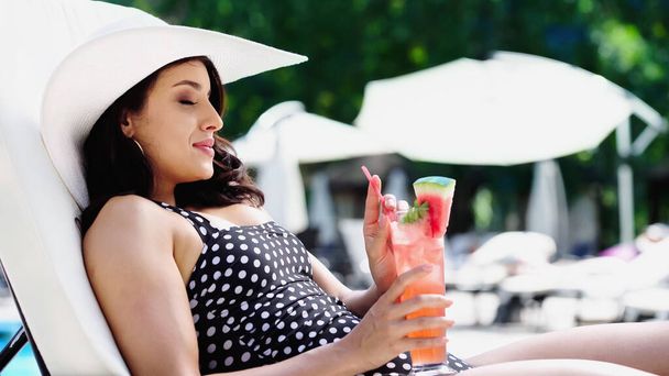 tevreden vrouw in gestippelde badpak en stro hoed drinken cocktail terwijl liggend op de ligstoel  - Foto, afbeelding