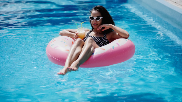 brünette Frau mit stylischer Sonnenbrille hält Glas mit Cocktail und schwimmt auf aufblasbarem Ring im Pool - Foto, Bild