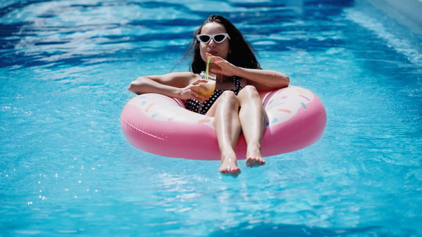 femme heureuse dans les lunettes de soleil élégantes boire cocktail et nager sur anneau gonflable dans la piscine - Photo, image