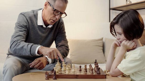 grandfather in glasses pointing at chess figure in living room - Fotó, kép