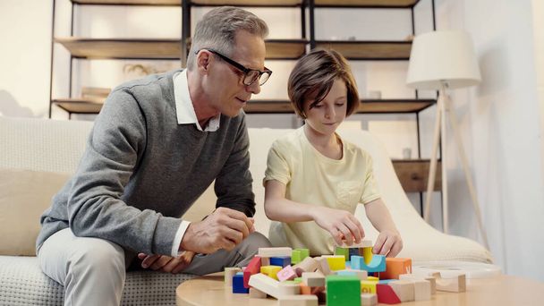 middle aged grandfather in glasses playing building blocks with preteen boy - Фото, изображение