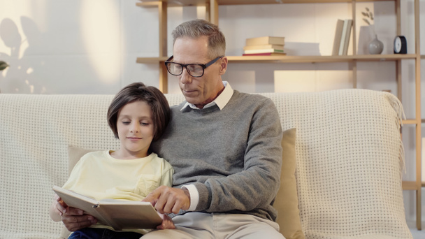 middle aged grandfather in glasses reading book with grandson at home - Photo, image