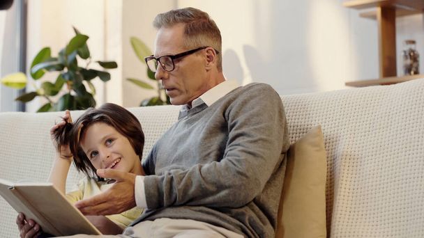 middle aged grandfather in glasses reading book with cheerful grandson at home - Foto, Bild