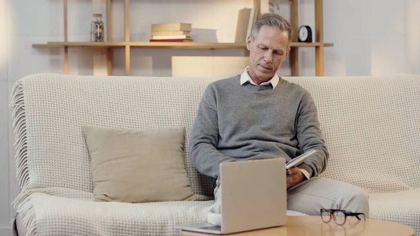 middle aged man reading book near laptop in living room - Fotoğraf, Görsel