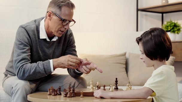 grandfather in glasses pointing at figure while playing chess with boy in living room - Photo, Image