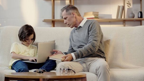 grand-père d'âge moyen tenant livre près de préadolescent petit-fils à l'aide d'un ordinateur portable dans le salon - Photo, image