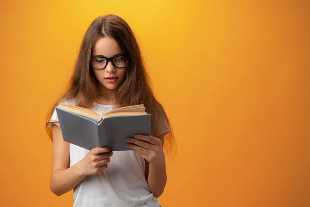 Smart teen girl in glasses holding book against yellow background - Zdjęcie, obraz