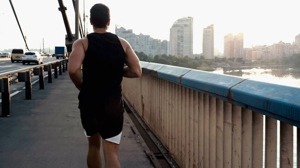 back view of athletic mixed race man running on city bridge over river - Photo, Image