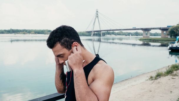 Atlétikai bi-rasszista férfi állító fülhallgató, miközben áll a folyó közelében - Fotó, kép