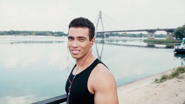 sport bi-razziale uomo in auricolare sorridente alla fotocamera vicino al fiume - Foto, immagini