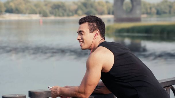 smiling mixed race man in earphones listening music on bridge over river - Photo, Image
