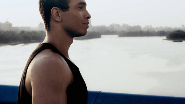 vista lateral del joven hombre bi-racial en el puente de la ciudad sobre el río - Foto, Imagen