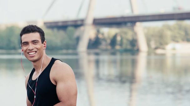 positive mixed race sportsman in earphones looking at camera near river and blurred bridge - Фото, изображение