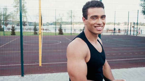 veselý bi-rasový sportovec při pohledu na kameru v blízkosti venkovní tělocvičny - Fotografie, Obrázek